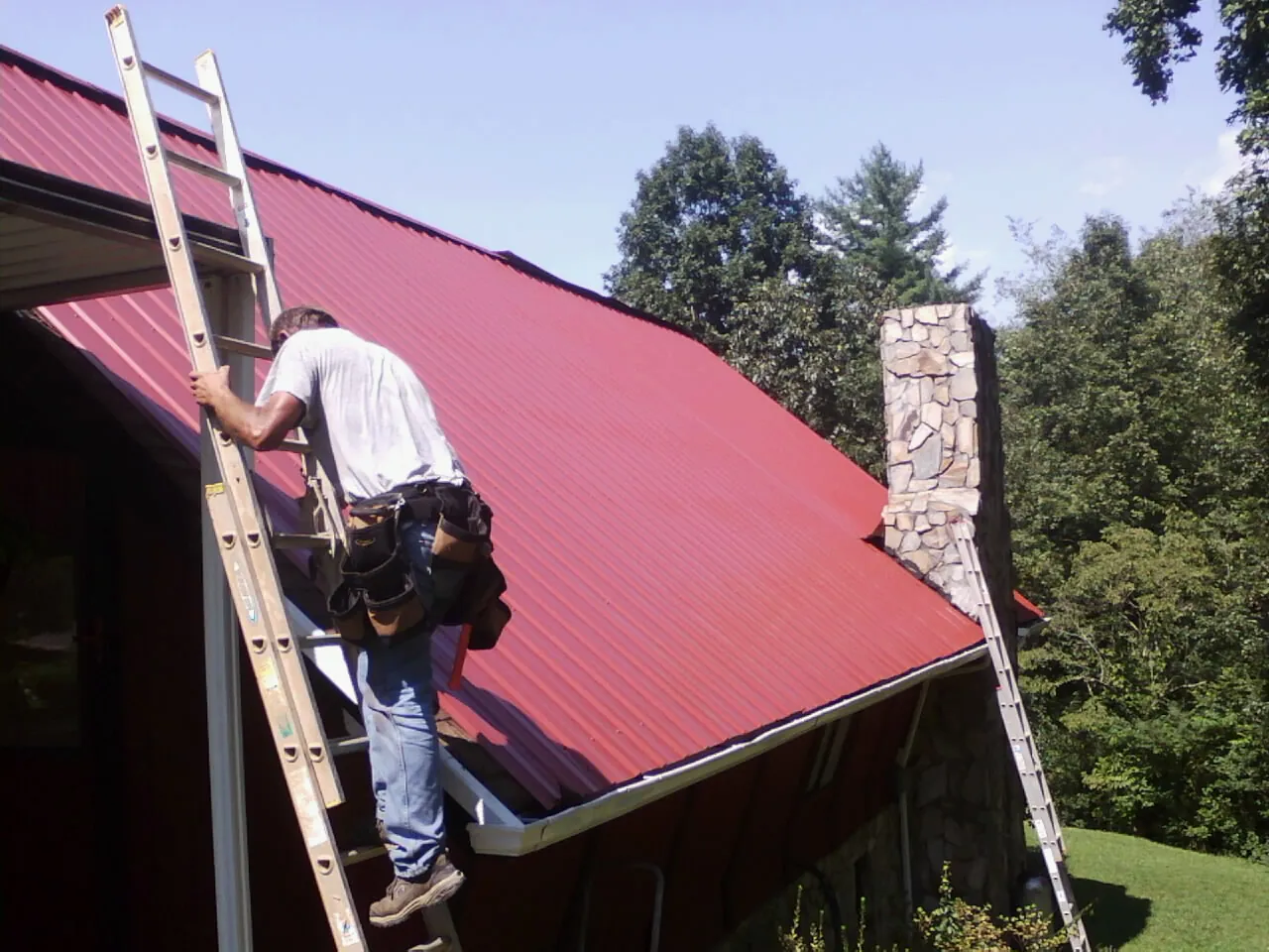 Roofer on a ladder beside a red metal roof and stone chimney — Soffit & Fascia Repair by Roofing Brothers in Green Pond